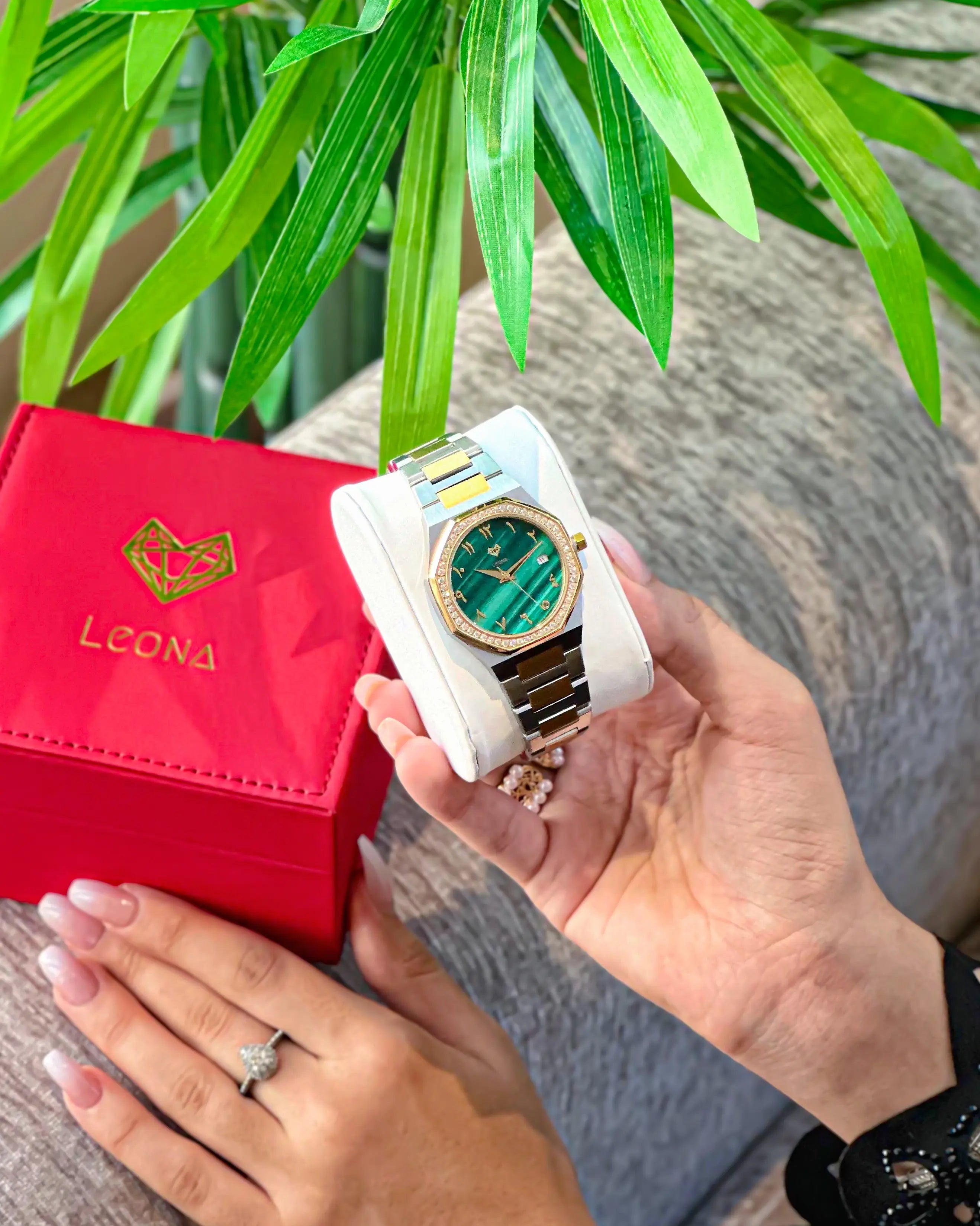 Person holding a white watch with a green face and gold accents, next to a red 'LEONA' box.