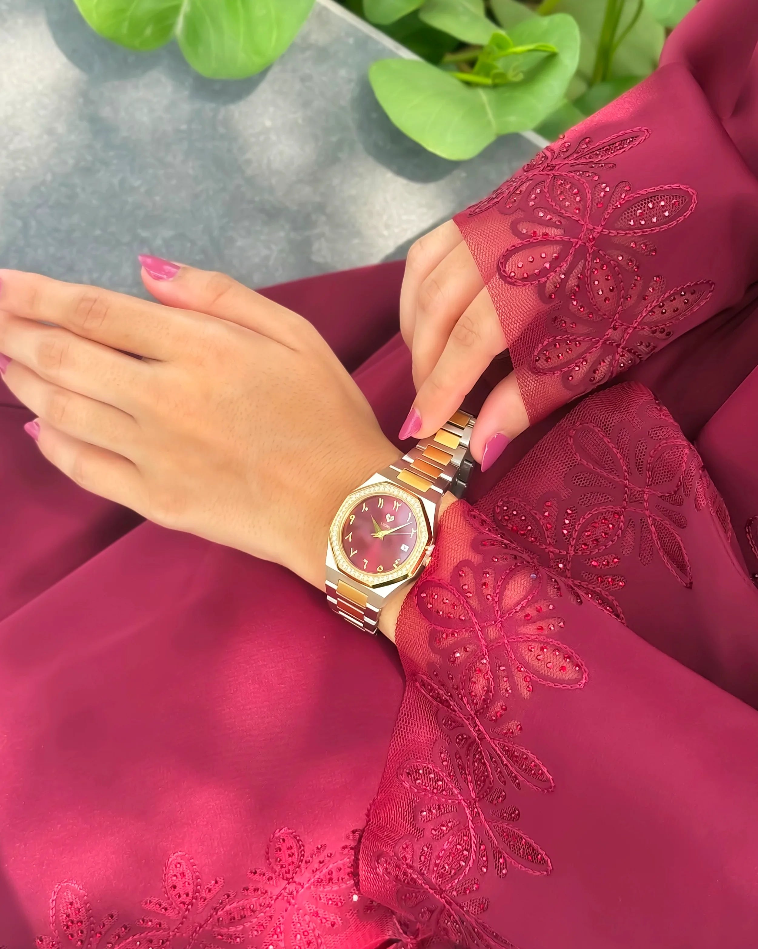Person wearing a pink and gold watch on a red embroidered garment with green leaves in the background