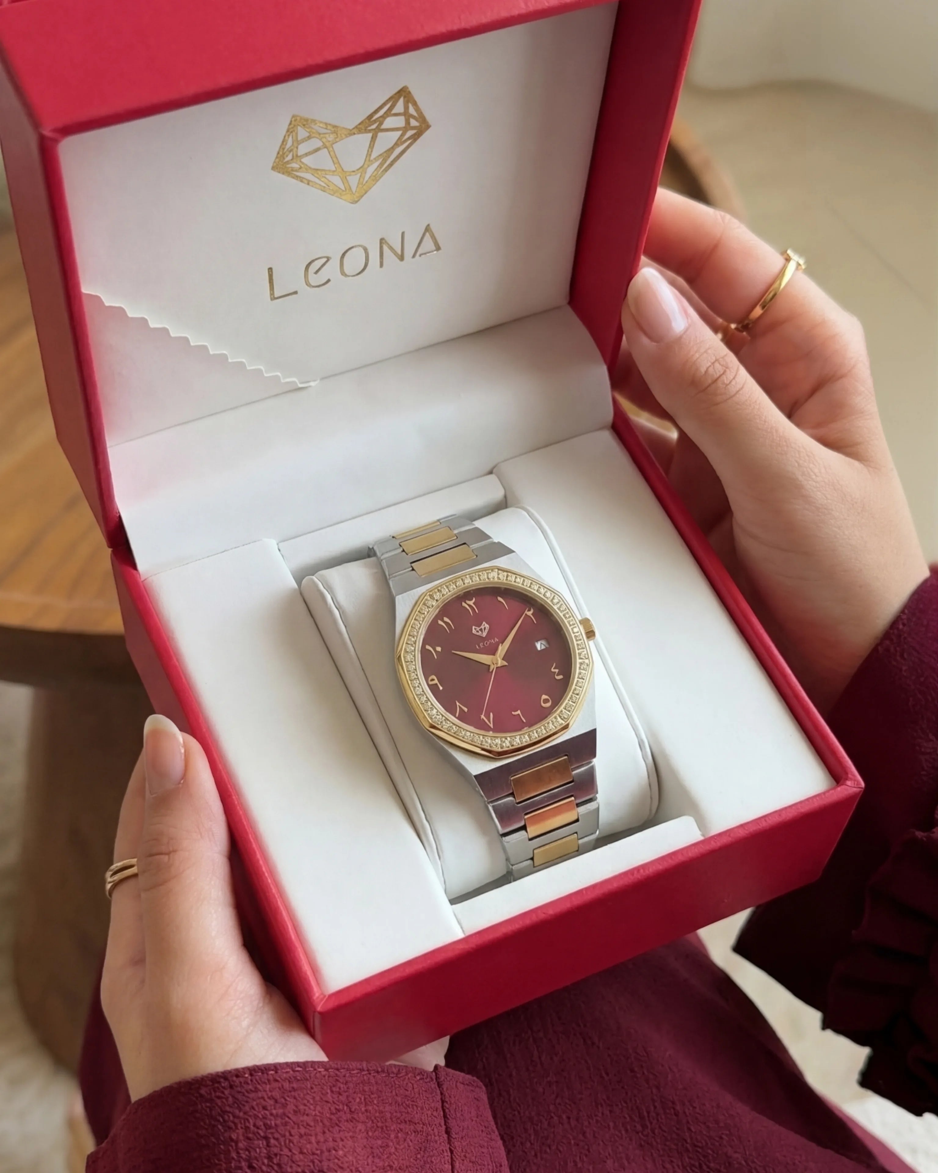 Watch in a red Leona box held by a person wearing a burgundy outfit.